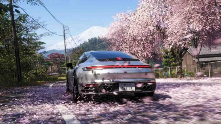 A rear-view shot of a silver Porsche 911 Carrera driving down a narrow road in Japan. The car is surrounded by falling pink cherry blossom petals that are blurred to suggest motion. To the right, large cherry blossom trees in full bloom hang over the road, while a snow-capped Mount Fuji looms in the distant background under a clear blue sky.