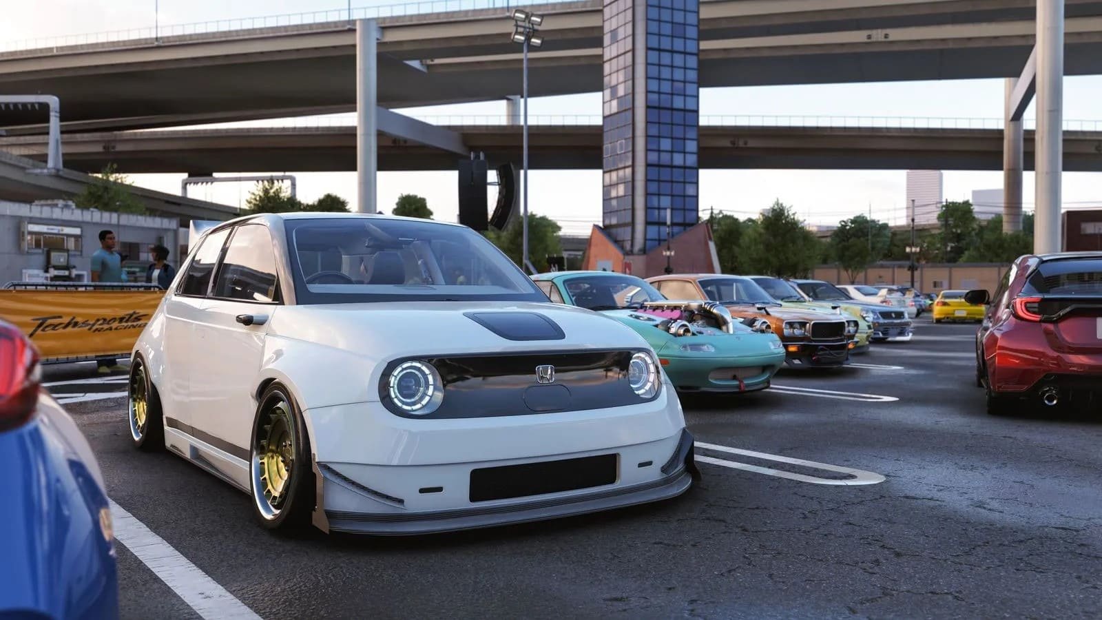 A wide-angle shot of a modified white Honda e electric car parked in an outdoor lot during a car meet. The car features a low stance, aftermarket gold-rimmed wheels, and an aggressive front splitter. In the background, other tuned Japanese cars and a large highway overpass are visible under a clear evening sky.