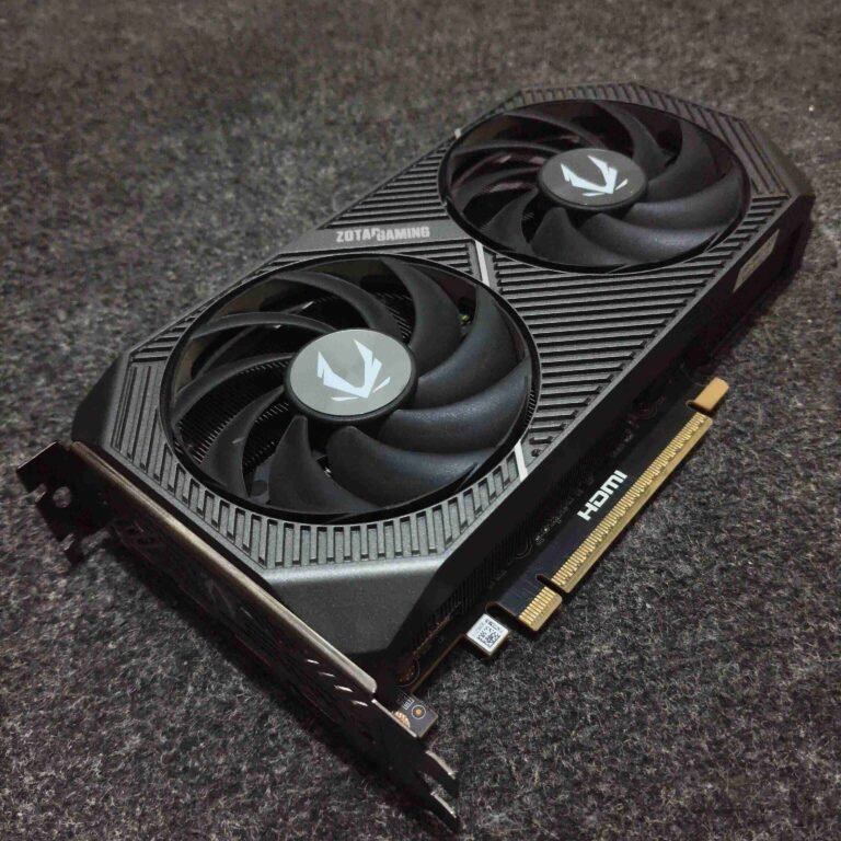 An angled, close-up shot of a ZOTAC GAMING Twin Edge OC GeForce RTX 5060 Ti 16GB graphics card resting on a dark, textured grey surface. The card features a sleek, compact black shroud with two large cooling fans, each sporting the silver ZOTAC logo in the center. The design includes diagonal ridges and the "ZOTAC GAMING" brand name visible near the top edge. The gold-plated PCIe connector and an HDMI port label are visible along the bottom edge of the hardware.