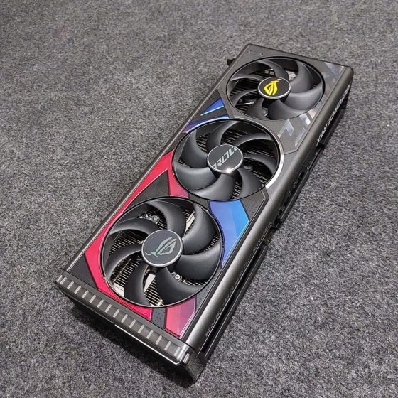 An overhead, angled shot of an ASUS ROG Strix GeForce RTX 4090 Gaming OC graphics card resting on a grey textured surface. The card features a massive, dark metallic industrial design with a triple-fan cooling system. The shroud is accented with a bold red and blue gradient geometric pattern around the fans. The center fan hub displays the "ROG" text, while the outer fans feature the Republic of Gamers "Eye" logo. A transparent acrylic strip with vertical ridges is visible at the end of the card, designed for RGB lighting.