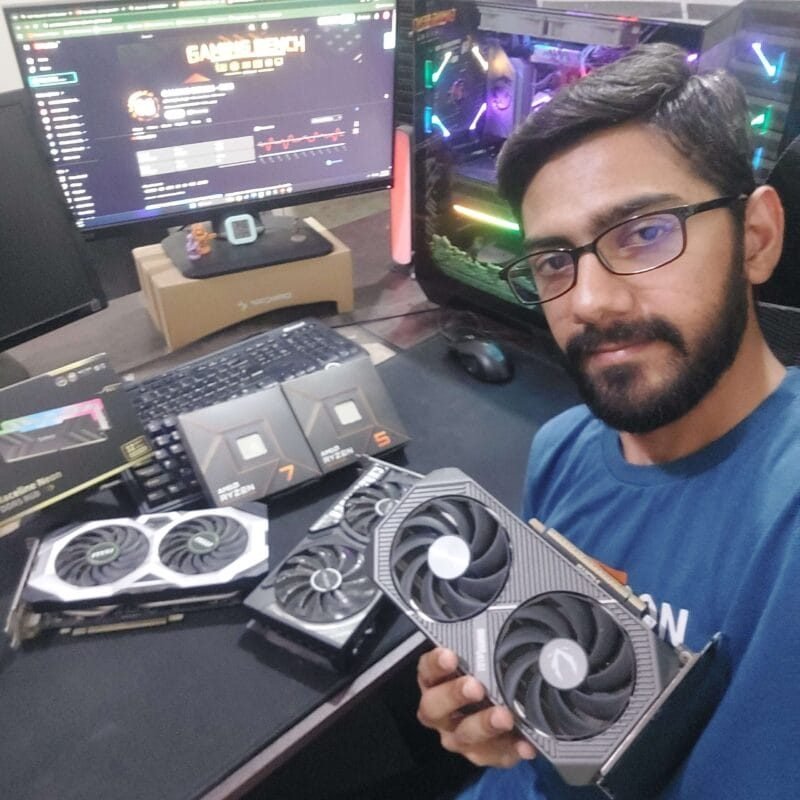 Shoaib Mughal, founder of Gaming Bench, holding a Zotac RTX 5060 Ti at his hardware testing desk with an RTX 5070 Ti system, CPUs, RAM, and GPUs visible in the background