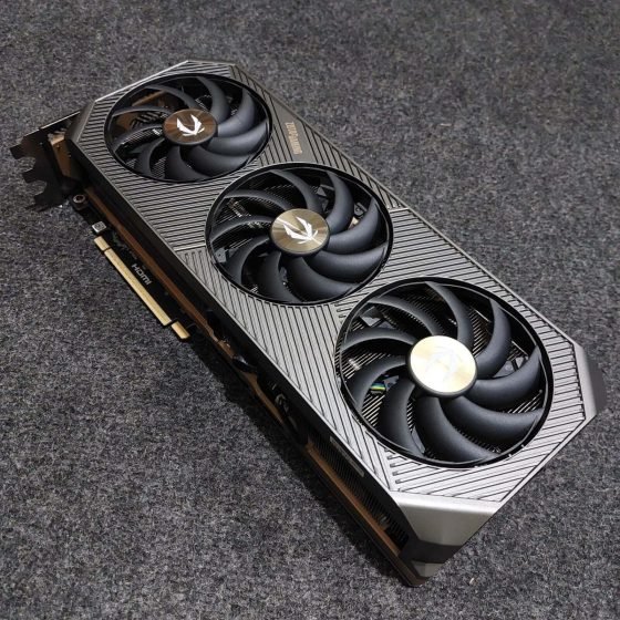 An angled, top-down studio shot of a ZOTAC GAMING GeForce RTX 5080 SOLID graphics card resting on a dark, textured grey surface. The card features a sleek, industrial design with a metallic grey shroud decorated with diagonal linear patterns. It is equipped with a triple-fan cooling system, featuring three large black fans with gold ZOTAC logos at the centers. The side of the card shows the PCIe connector, an HDMI port, and a glimpse of the thick internal heatsink fins.